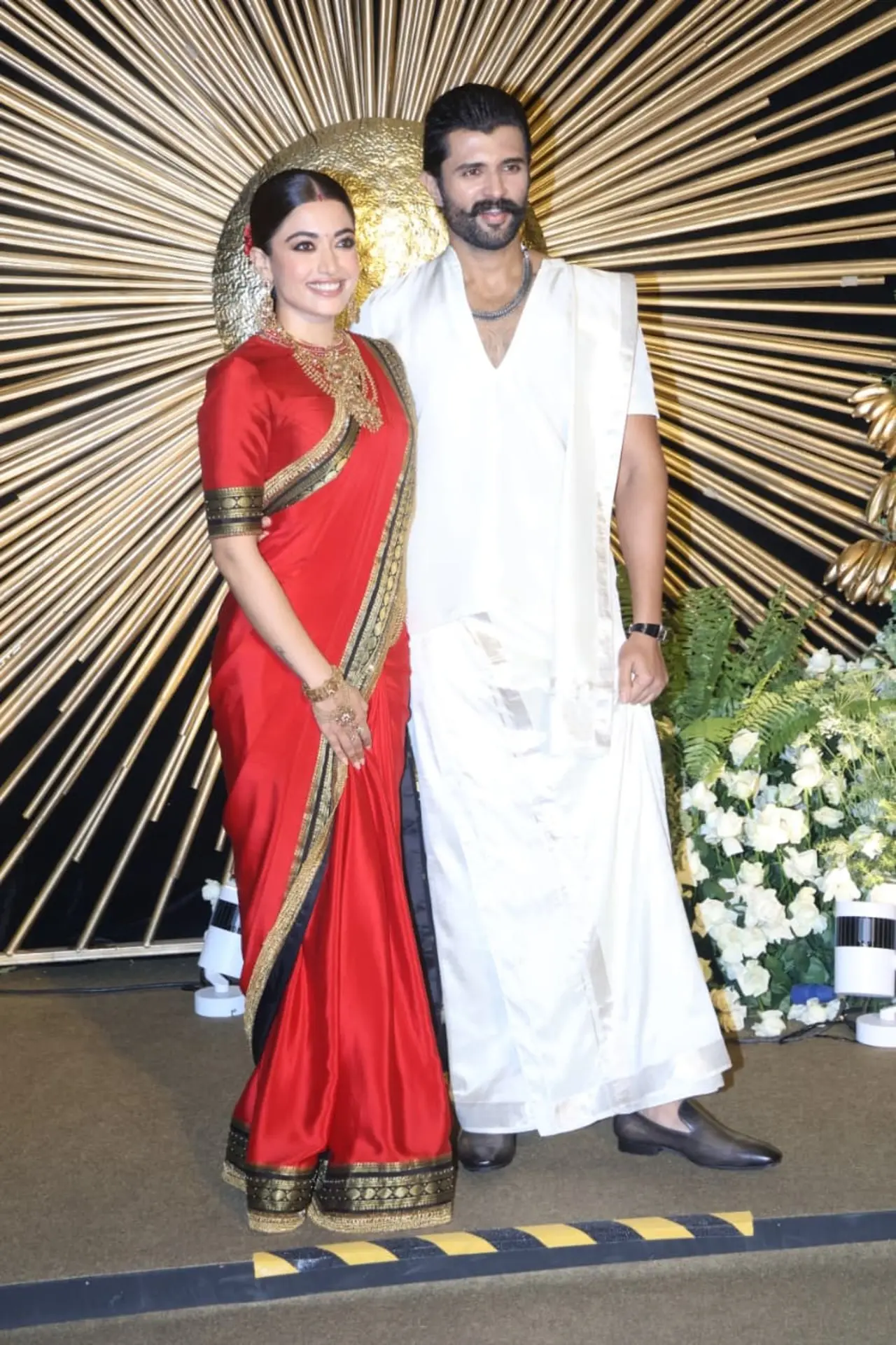 Rashmika Mandanna wore a red silk saree with a black-and-gold border for her reception. She paired it with heavy gold temple jewellery. Vijay complements Rashmika in traditional ensemble in a simple yet classic white veshti. He accessorises with layered metallic chains around his neck.